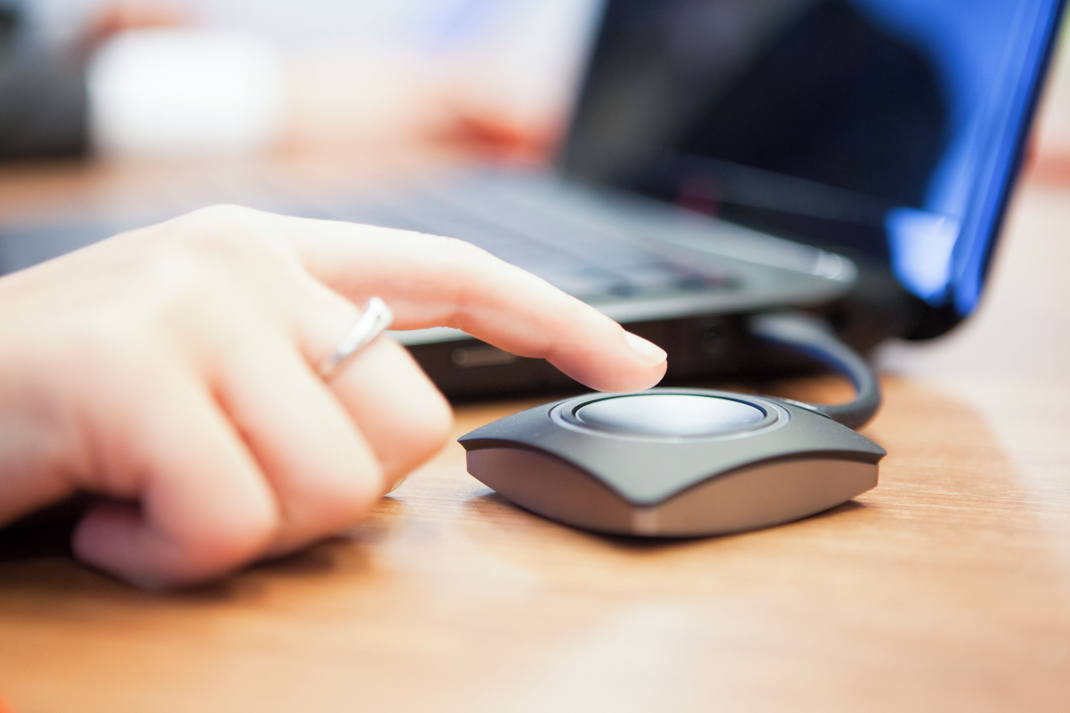 A hand using a modern, compact mouse on a wooden desk with a laptop in the background.
