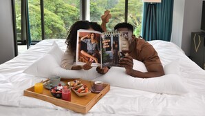 A couple lying on a bed with a magazine, a tray with breakfast, and a window with a view of trees. - Novotel Nairobi Westlands (Nairobi)