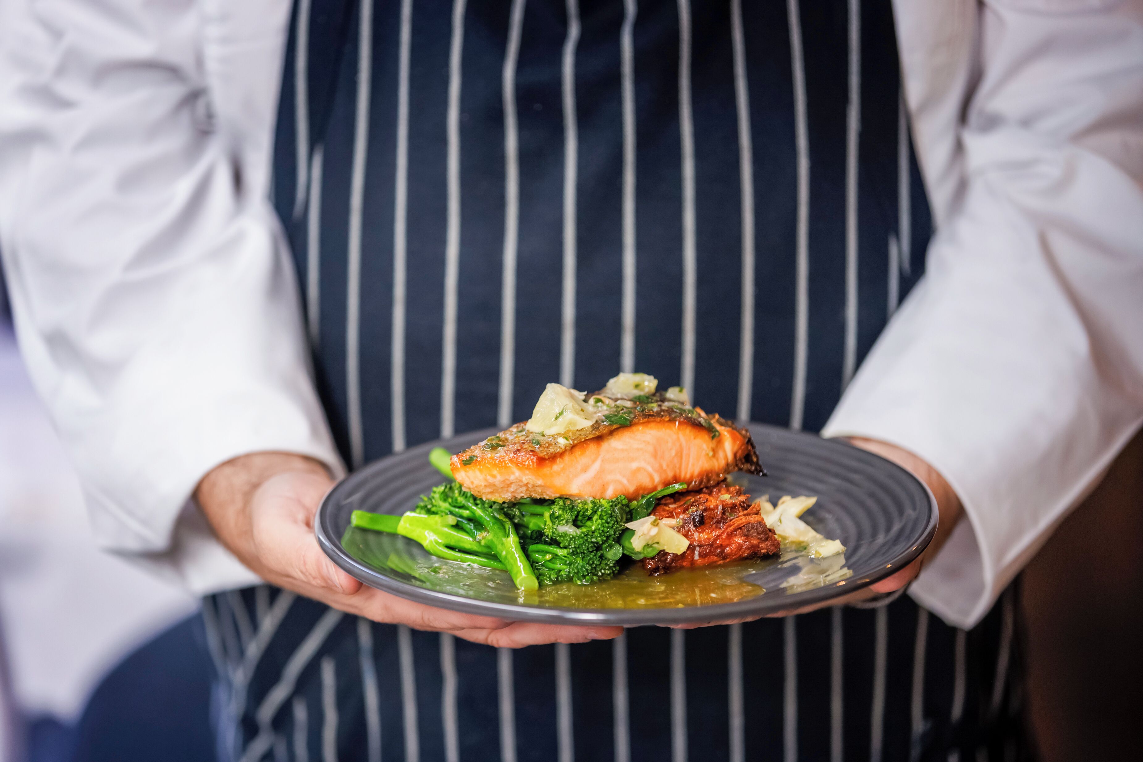 A person holding a plate with salmon, broccoli, and a side dish.