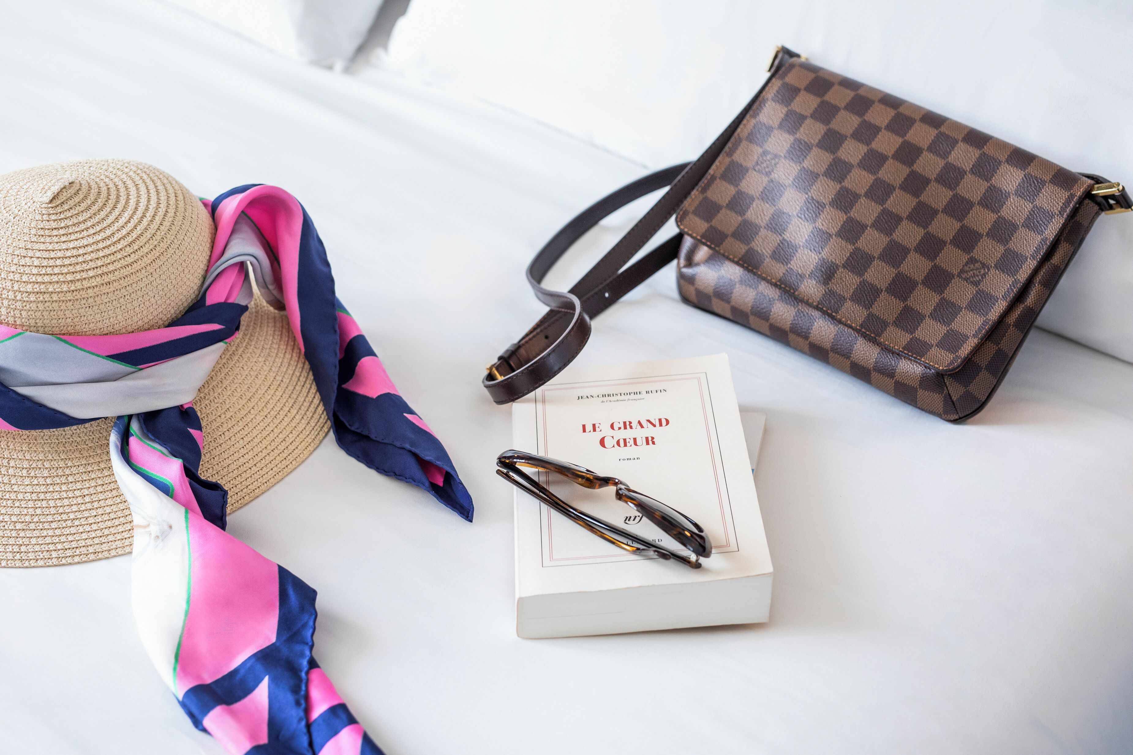A bed with a straw hat, a colorful scarf, a checkered handbag, a pair of glasses, and a book titled "Le Grand Cœur".