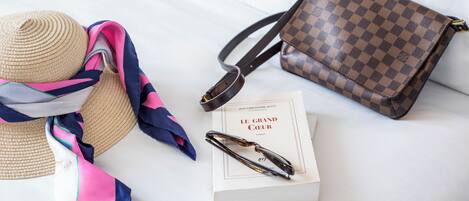 A bed with a straw hat, a colorful scarf, a checkered handbag, a pair of glasses, and a book titled "Le Grand Cœur".