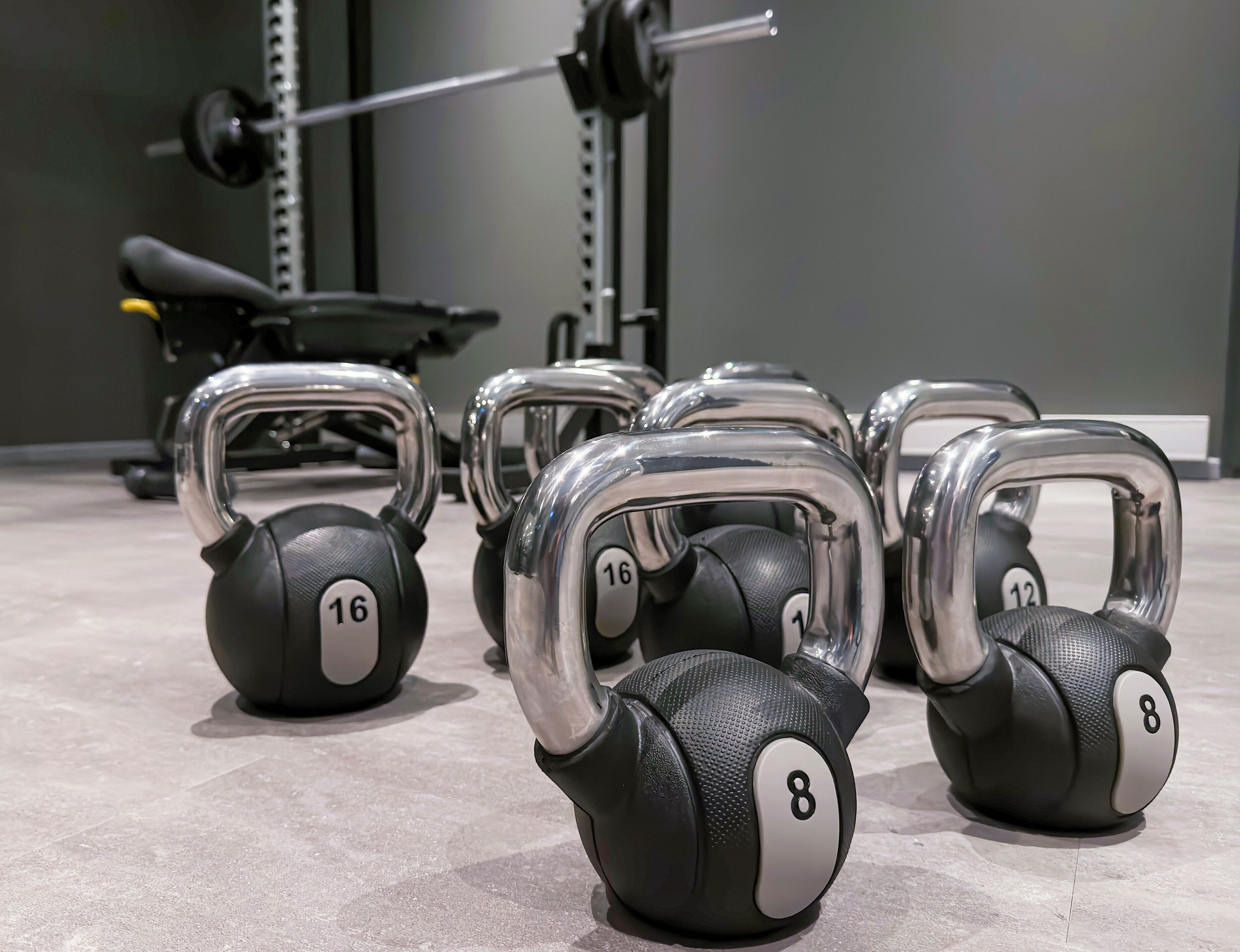 A set of kettlebells with weights of 8 and 16 kg, arranged on a gym floor with a barbell and weight rack in the background.