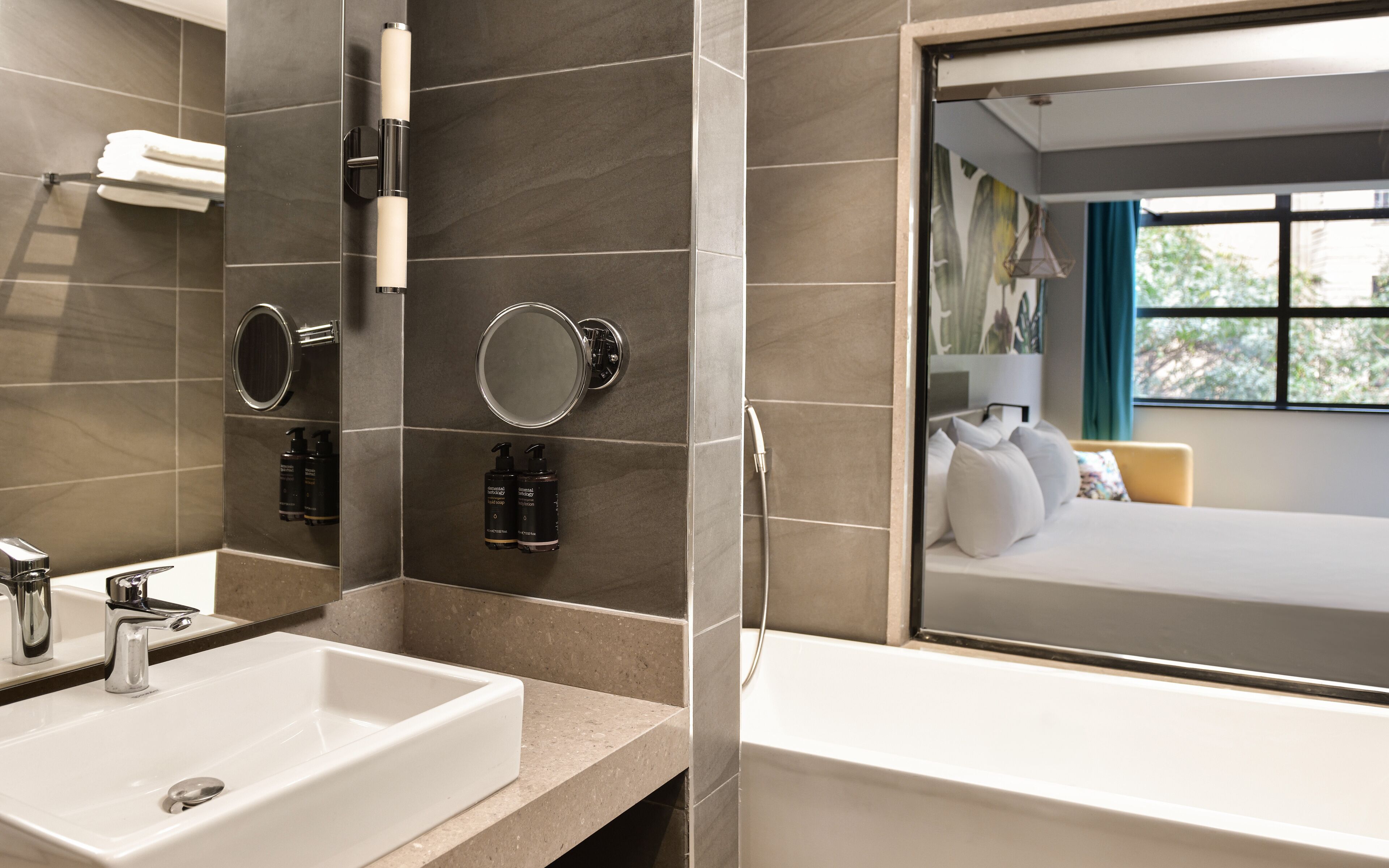 Un baño moderno con dos lavamanos, un espejo y artículos de aseo. Una ventana muestra un dormitorio con una cama y vistas a zonas verdes.
