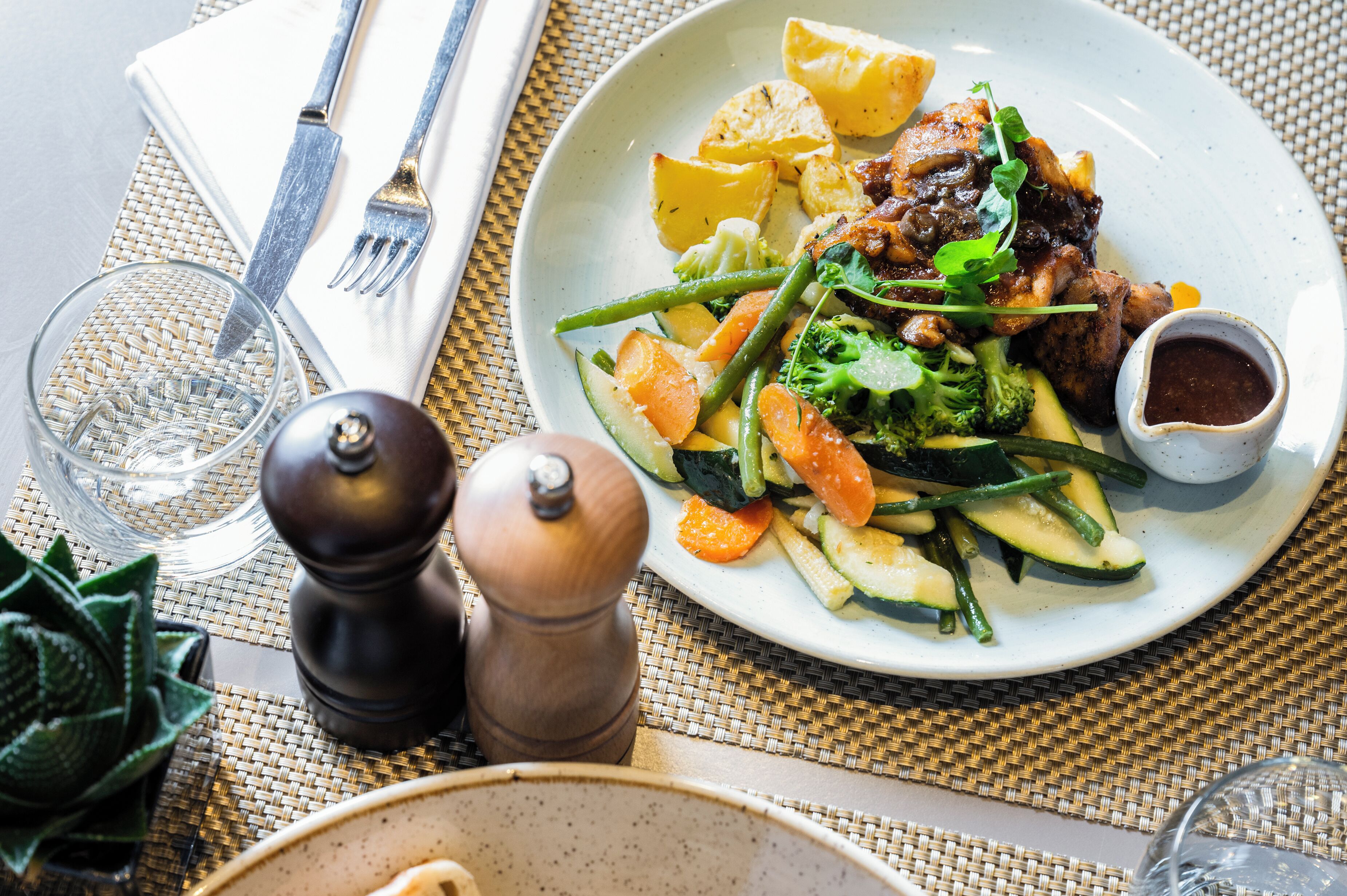 A plate of food with vegetables and meat, a glass of water, and a pepper grinder on a table.