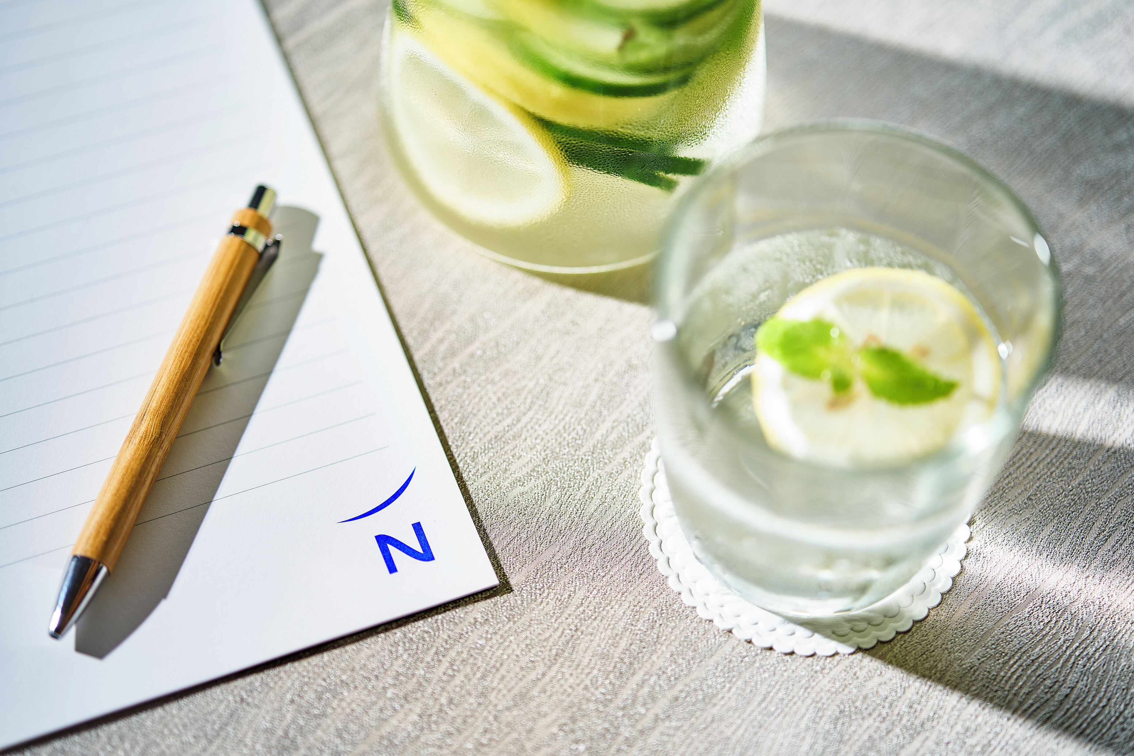 En notesblok med en pen, et glas vand med citronskiver, og en vandflaske på en struktureret overflade.