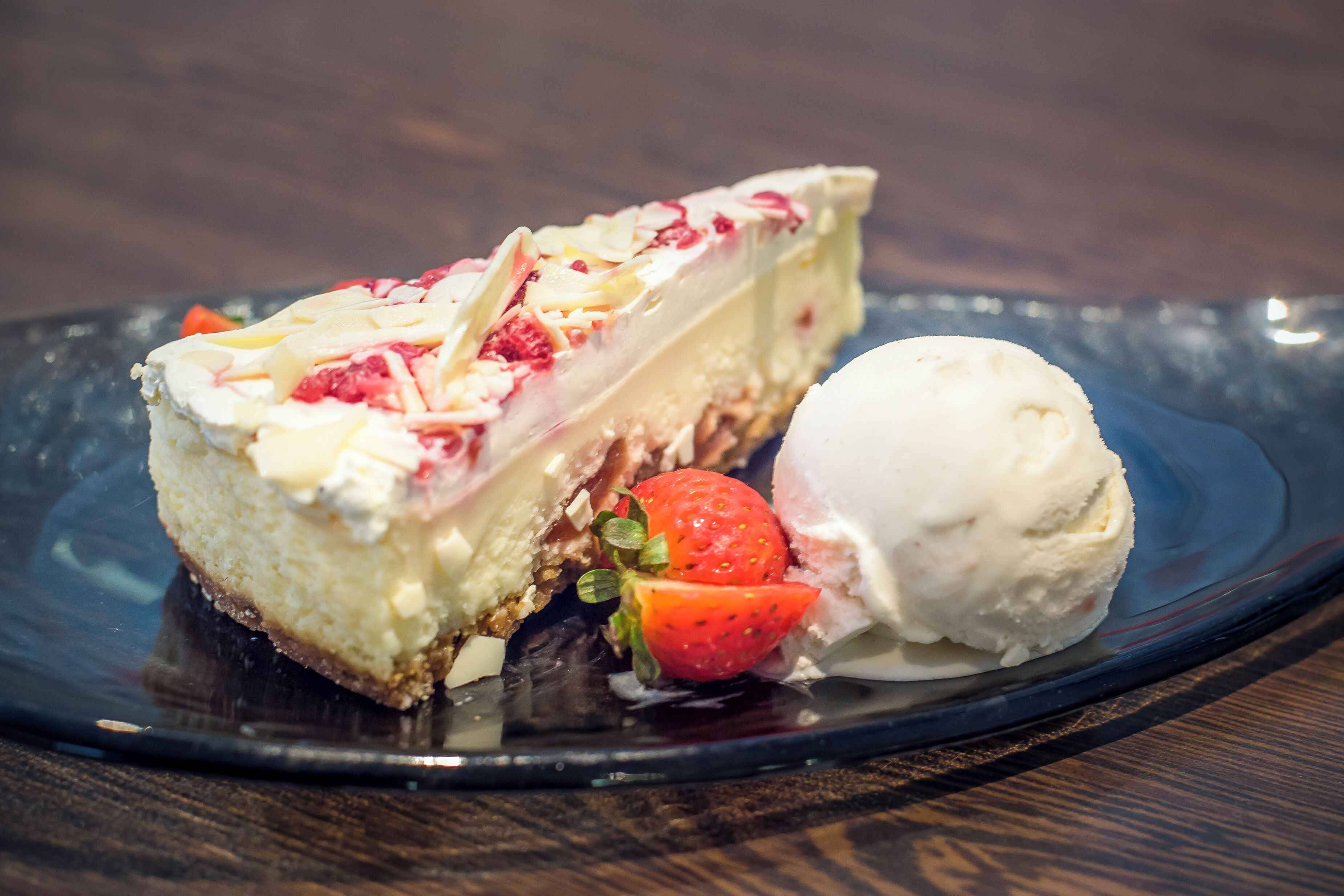 A slice of cheesecake with strawberries and a scoop of vanilla ice cream on a dark plate.
