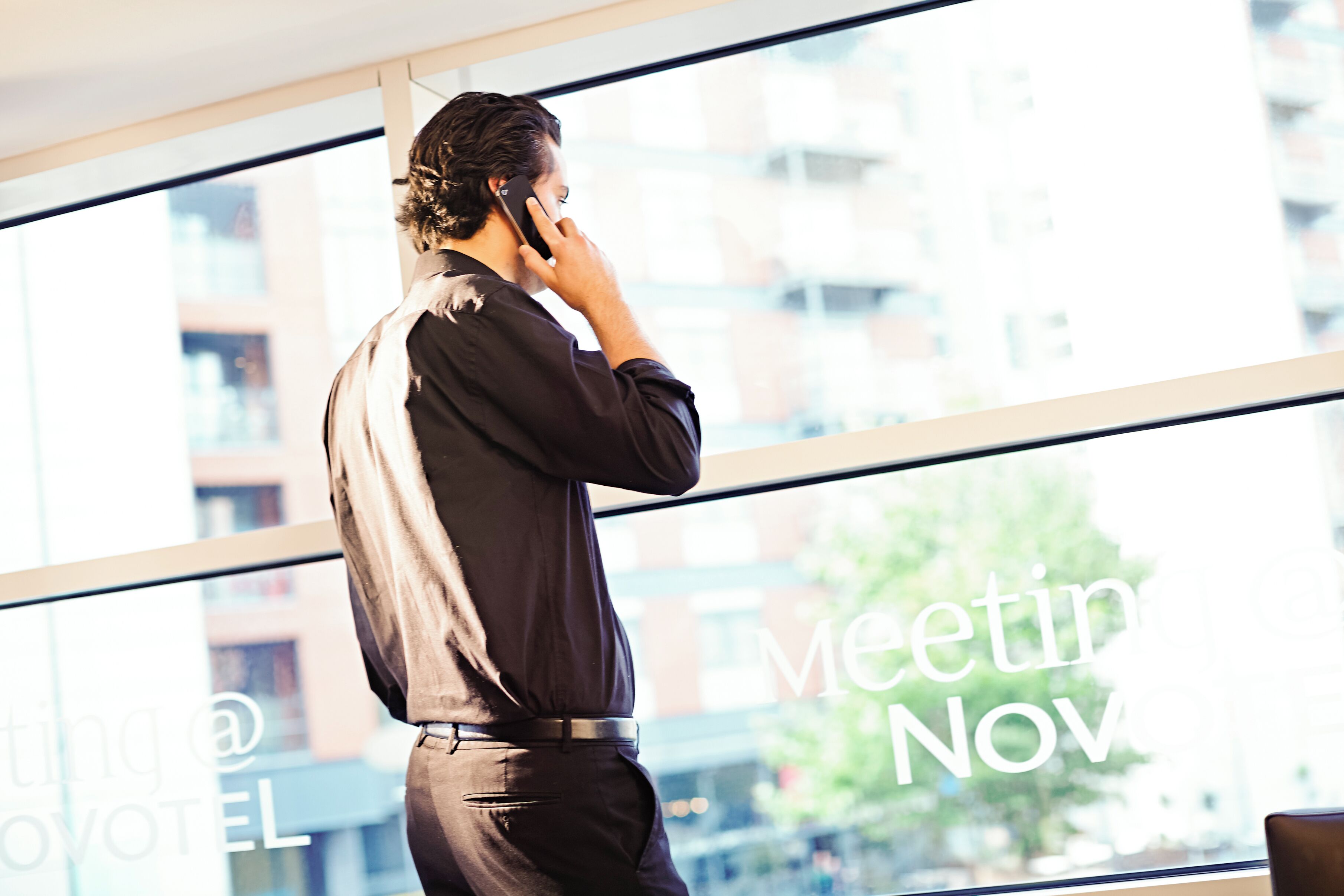 Eine Person im Business-Anzug steht an einem Fenster mit Blick auf die Stadt und telefoniert.