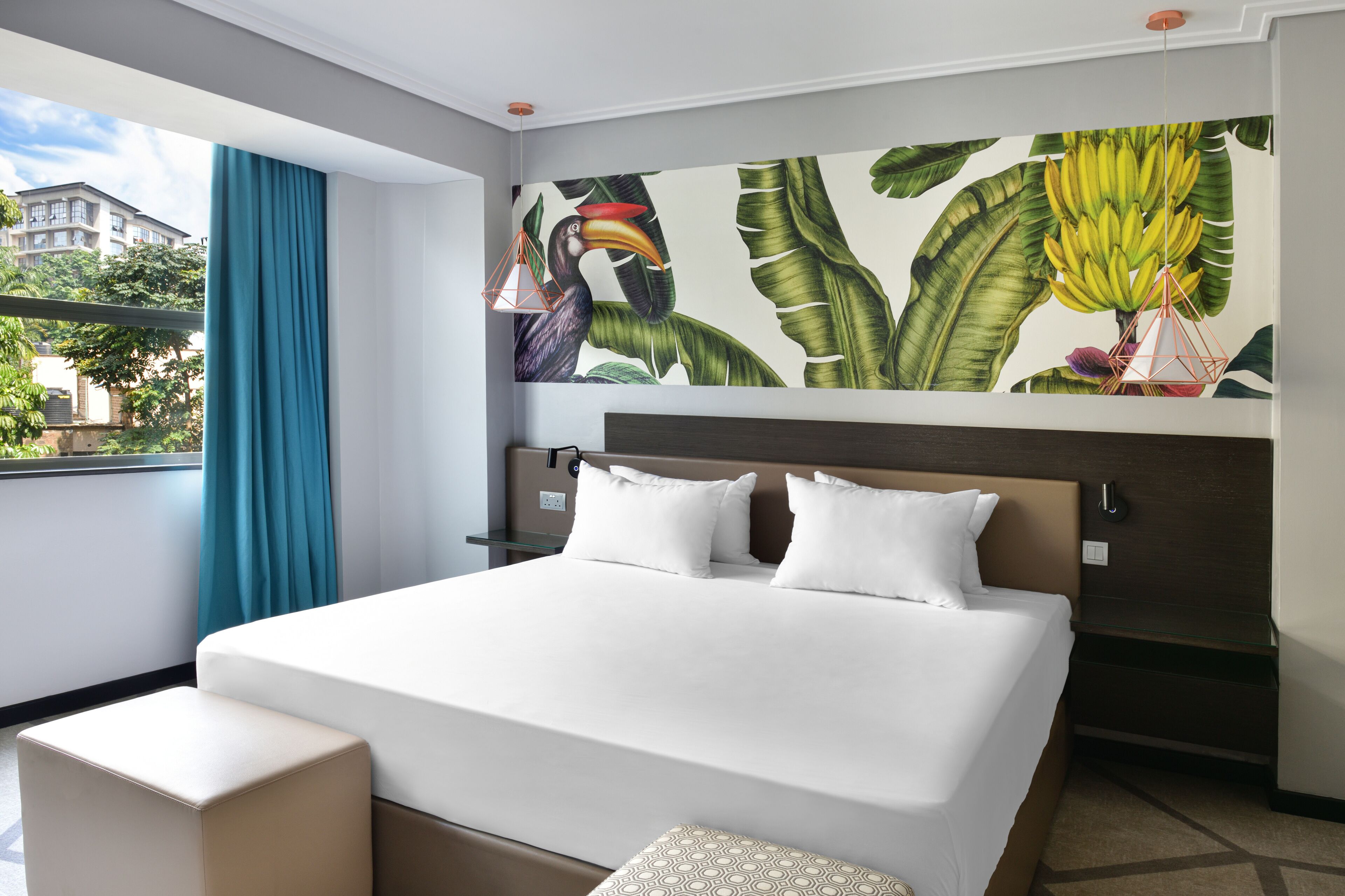 Habitació d'hotel amb un llit gran, una finestra amb cortines, un mural a la paret amb fulles tropicals i un aguiló, i vista a edificis a l'exterior.
