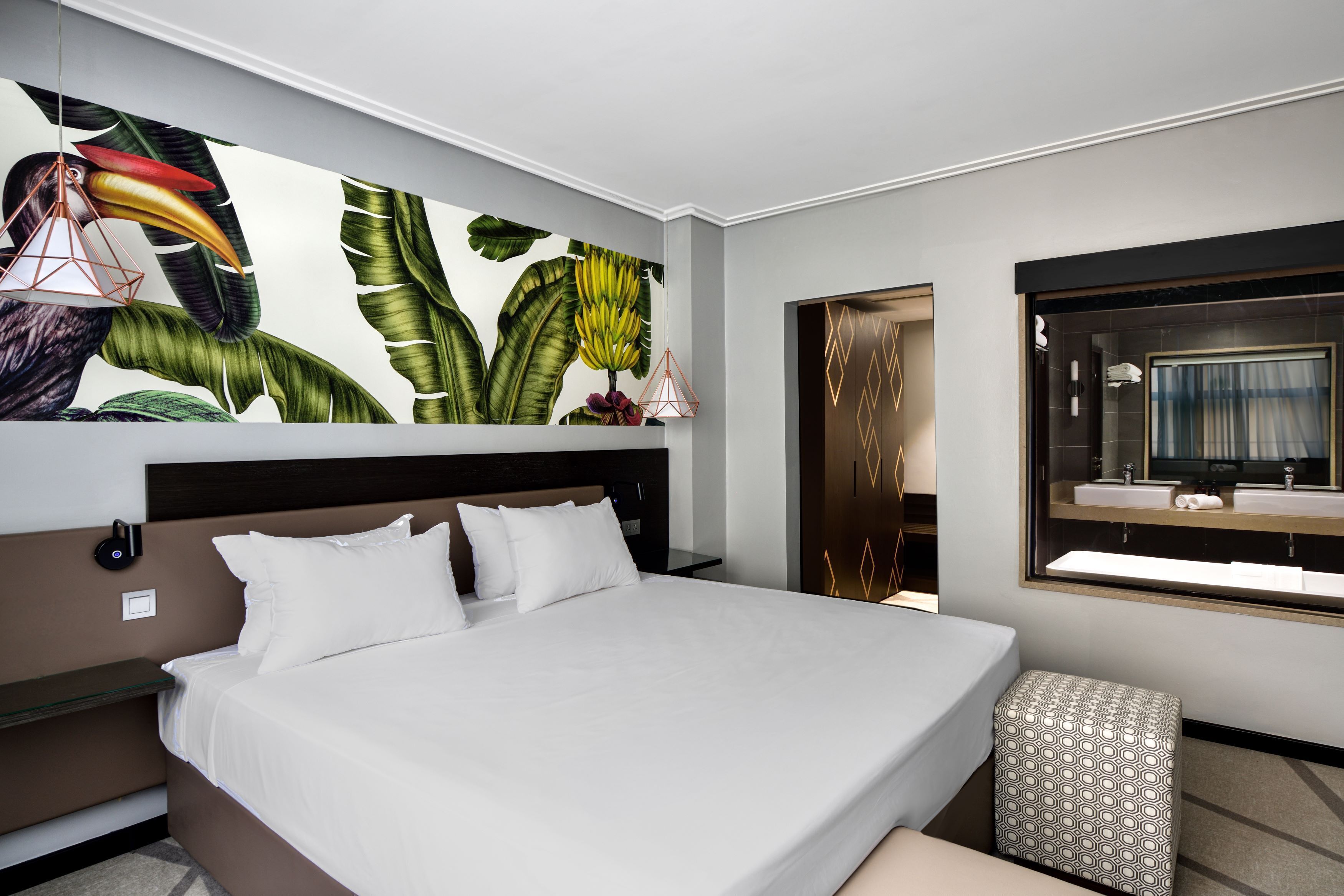 Une chambre d’hôtel moderne avec un grand lit, une fresque murale représentant des feuilles tropicales, une table de chevet avec une lampe et une salle de bain dotée d’un grand miroir et d’un lavabo.