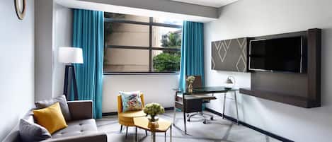 A modern hotel room with a grey sofa, a small table with a vase, a desk, a chair, and a mounted TV.
