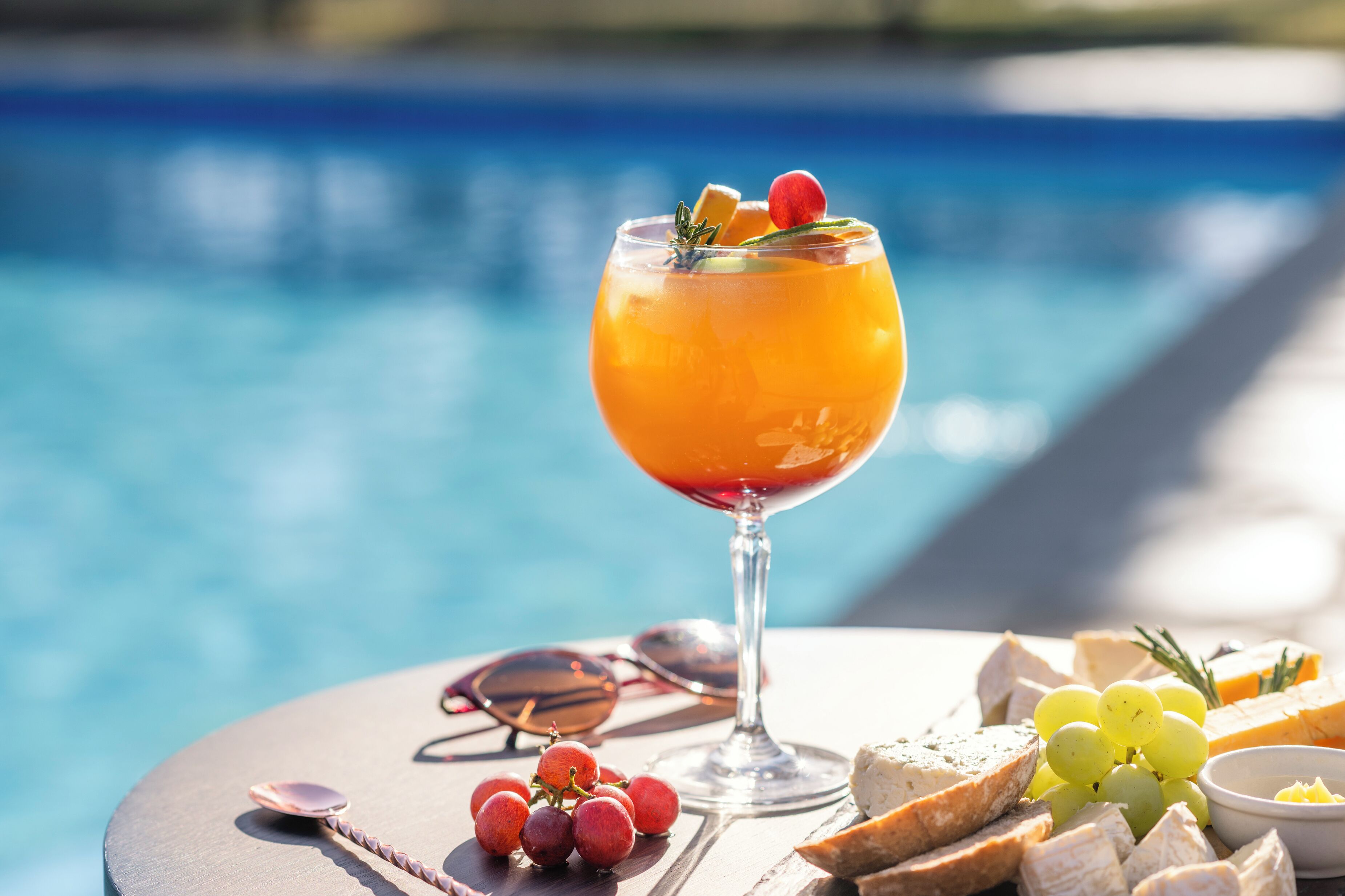 A table with a glass of orange drink, grapes, cheese, and bread by a pool.