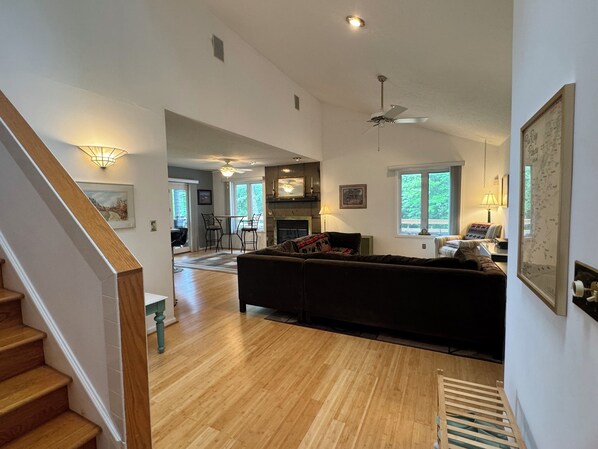 A spacious living room with hardwood floors, a dark-colored sofa, a dining area with a table and chairs, and a staircase with wooden steps.
