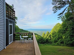 A wooden deck with white chairs and a table, overlooking a lush green landscape and distant mountains.