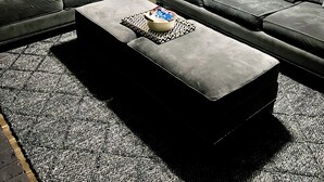 Sala de estar com sofá cinza, um banquinho combinando e uma mesa de centro com uma tigela de petiscos. Uma porta e uma cozinha aparecem ao fundo.