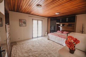 A bedroom with a wooden ceiling, a bed with a pink blanket, a dresser, and a window with blinds. - Portal Flores Suítes (Campos do Jordão)