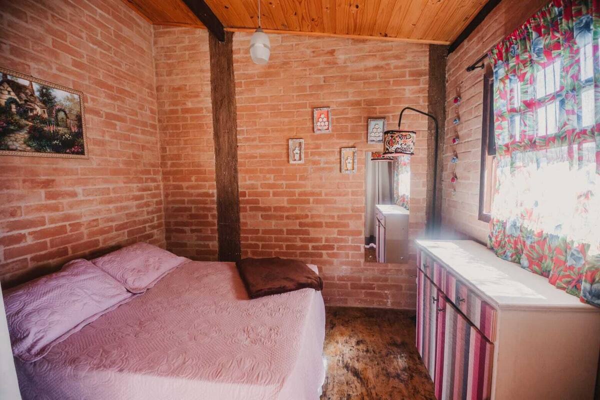 Une pièce avec un lit, une commode, une fenêtre avec des rideaux et un mur en briques.