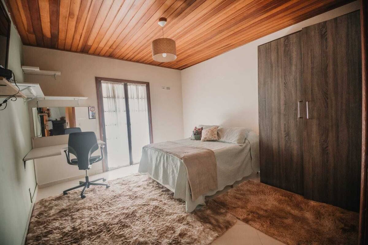 Une chambre avec un plafond en bois, un lit recouvert d’une couvre-lit de couleur claire, un bureau avec une chaise et une armoire.