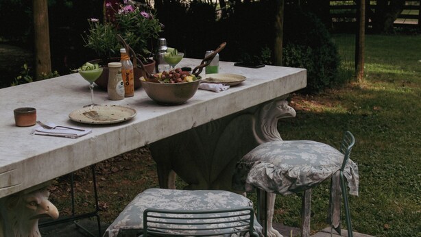 A stone table with a bowl of fruit, a bottle, and a glass. There are two metal chairs with cushions. The setting is outdoors with grass and trees in the background.
