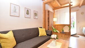 A cozy living room with a grey sofa, yellow cushions, and a wooden coffee table with a vase of flowers. There are framed botanical prints on the wall and a wooden ceiling. - Ferienwohnung in Rerik bis zu 2 Personen (Rerik (Ostseebad))