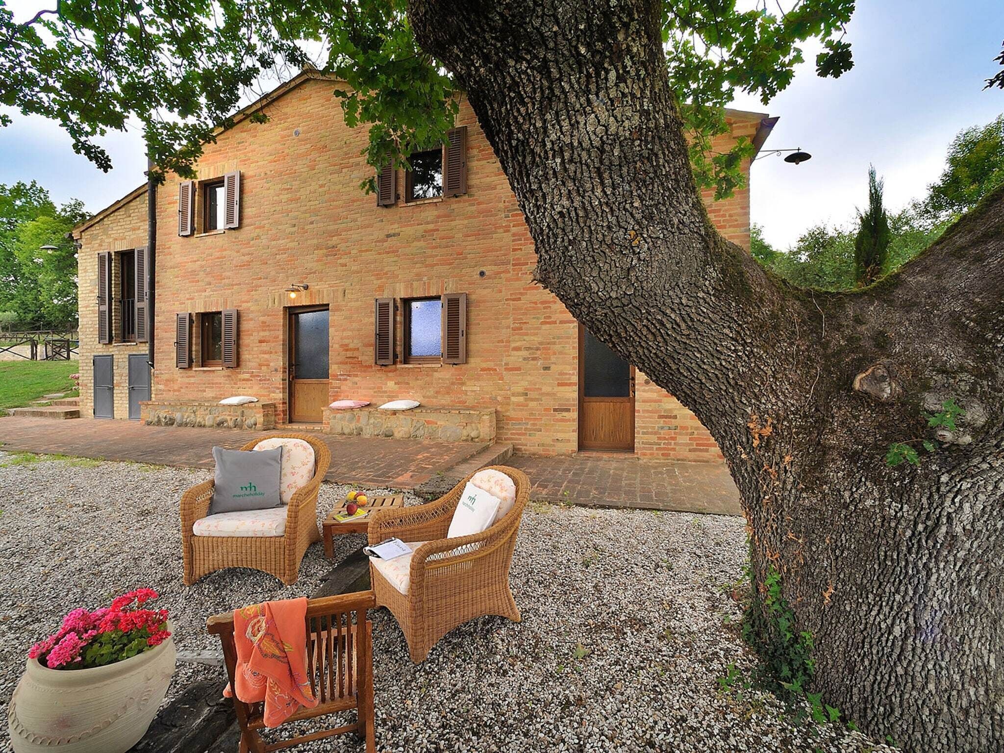 Una casa rural de maó amb un pati de grava, mobles de vimbles i un gran arbre.