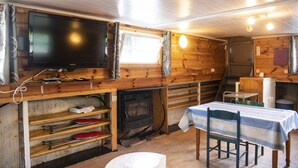 A cabin interior with a television, wooden walls, a table with chairs, a fireplace, and a window with curtains. - Idyllic Lakeside Cottage (Florennes)