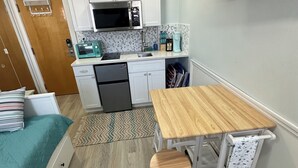 Cocina compacta con armarios blancos, microondas y cafetera. Hay una pequeña mesa de madera con una silla, una cama con ropa de cama color turquesa, y un televisor montado en la pared.