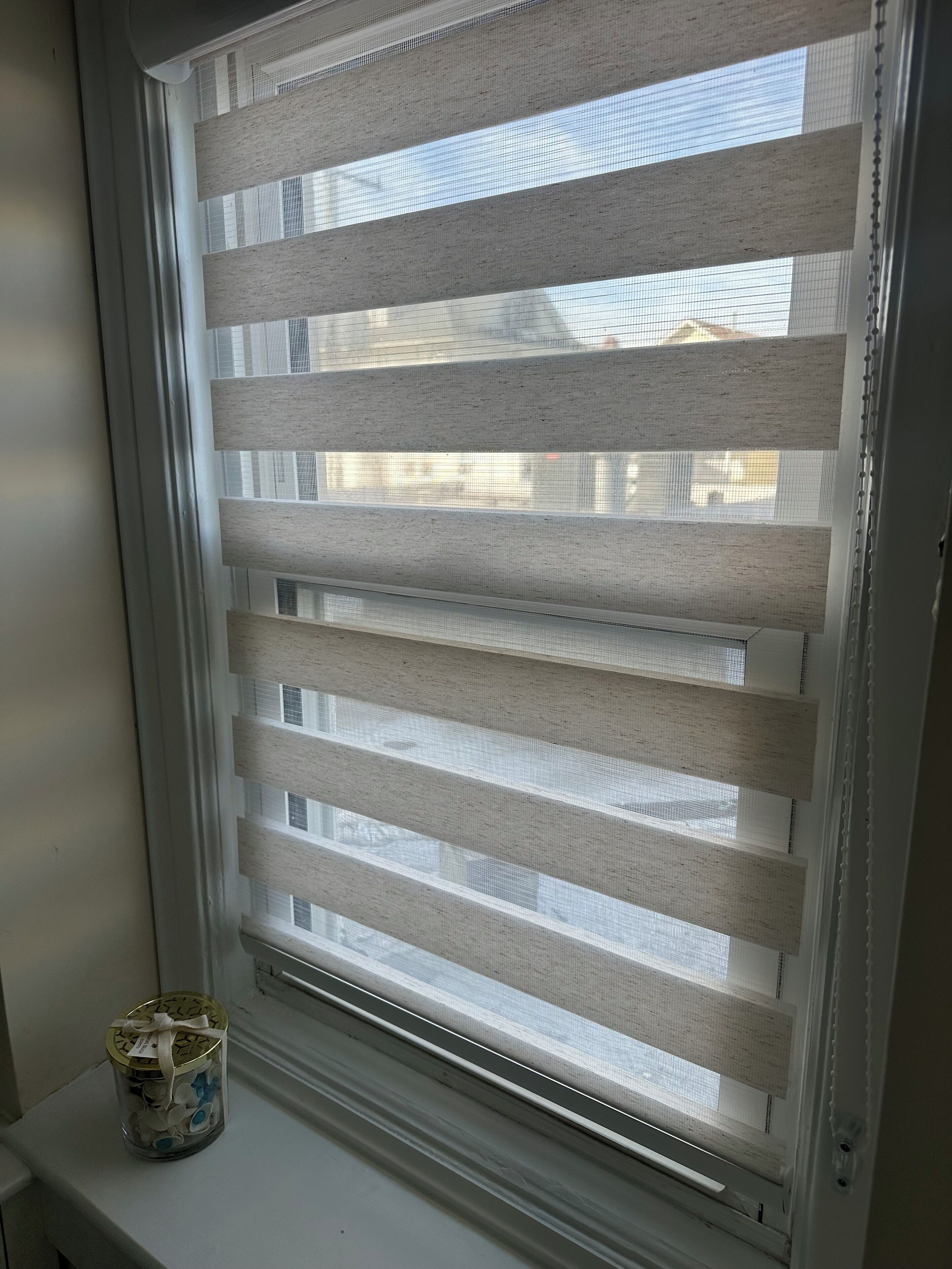 A window with horizontal blinds, a small decorative jar on the sill, and a view of buildings outside.