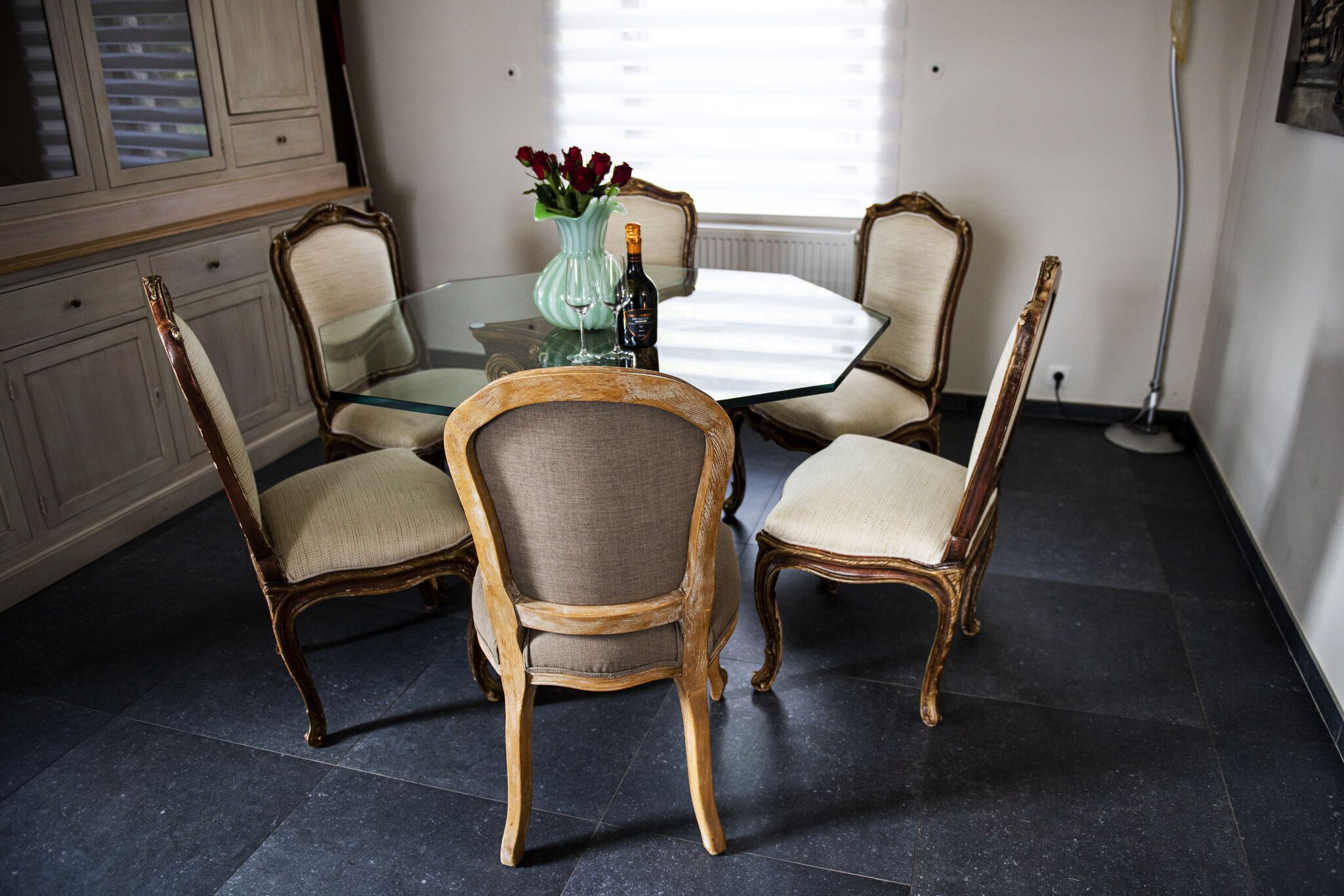A dining area with a glass table surrounded by four wooden chairs with cushioned seats. A vase with red flowers and a bottle of wine are on the table.