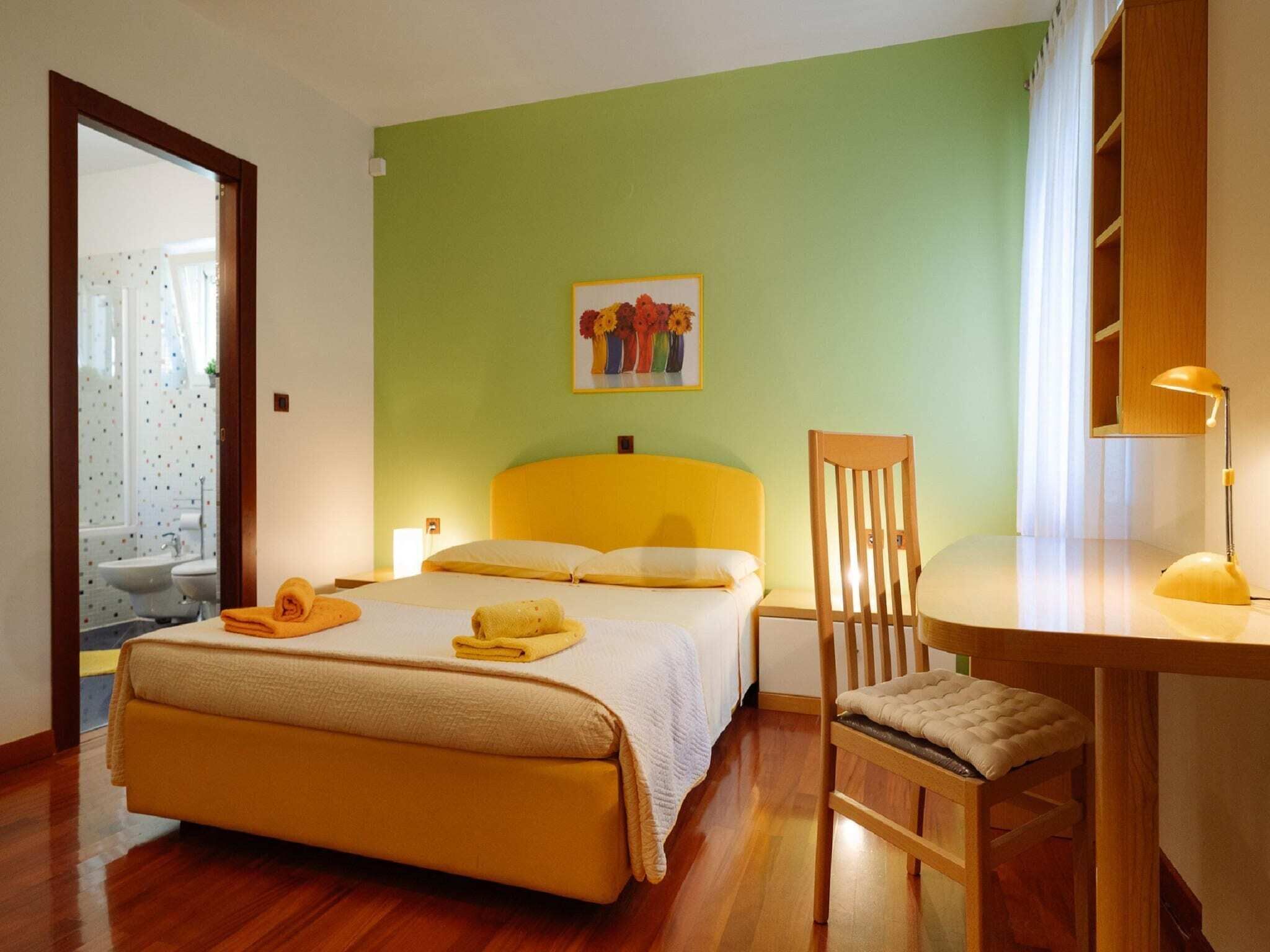 Une chambre d’hôtel avec un lit, un bureau et une chaise, un tableau accroché au mur et une salle de bain visible à travers une porte ouverte.