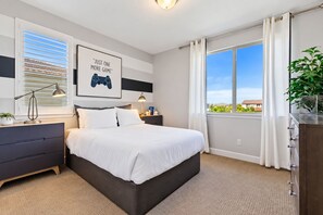 A bedroom with a bed, bedside tables, a desk, a lamp, a window with curtains, and a wall-mounted picture. - Blue Heaven (Roseville)