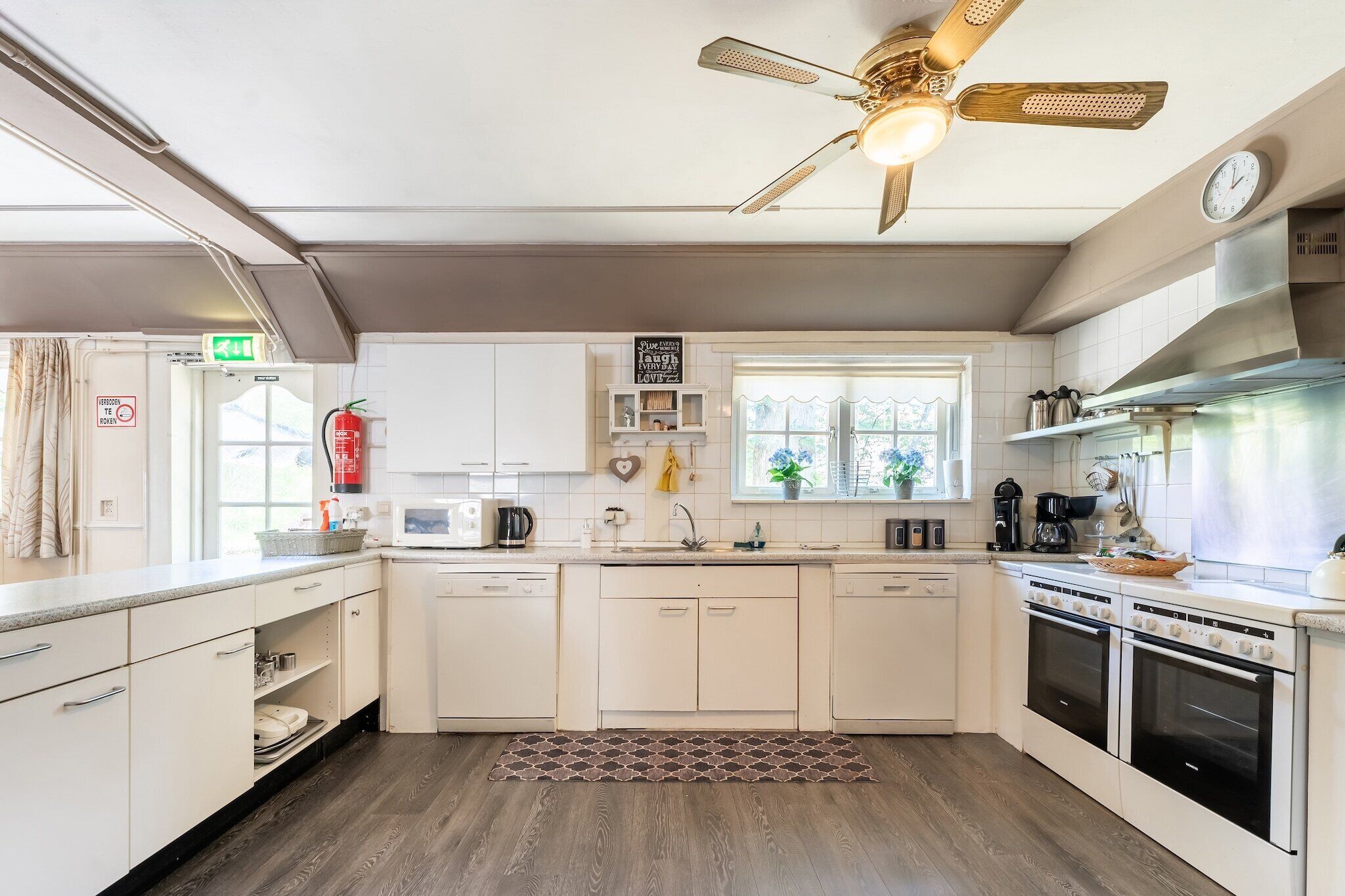 A modern kitchen with white cabinets, a microwave, a sink, and a stove. There is a ceiling fan, a clock, and a window with plants.