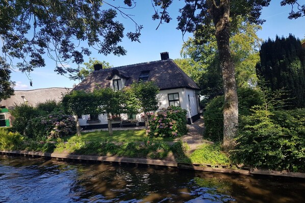 A house by a canal with a thatched roof, surrounded by greenery and a wooden bridge.