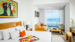 Habitación de hotel con una cama grande, un sofá, una mesa de comedor con desayuno y vista al océano.