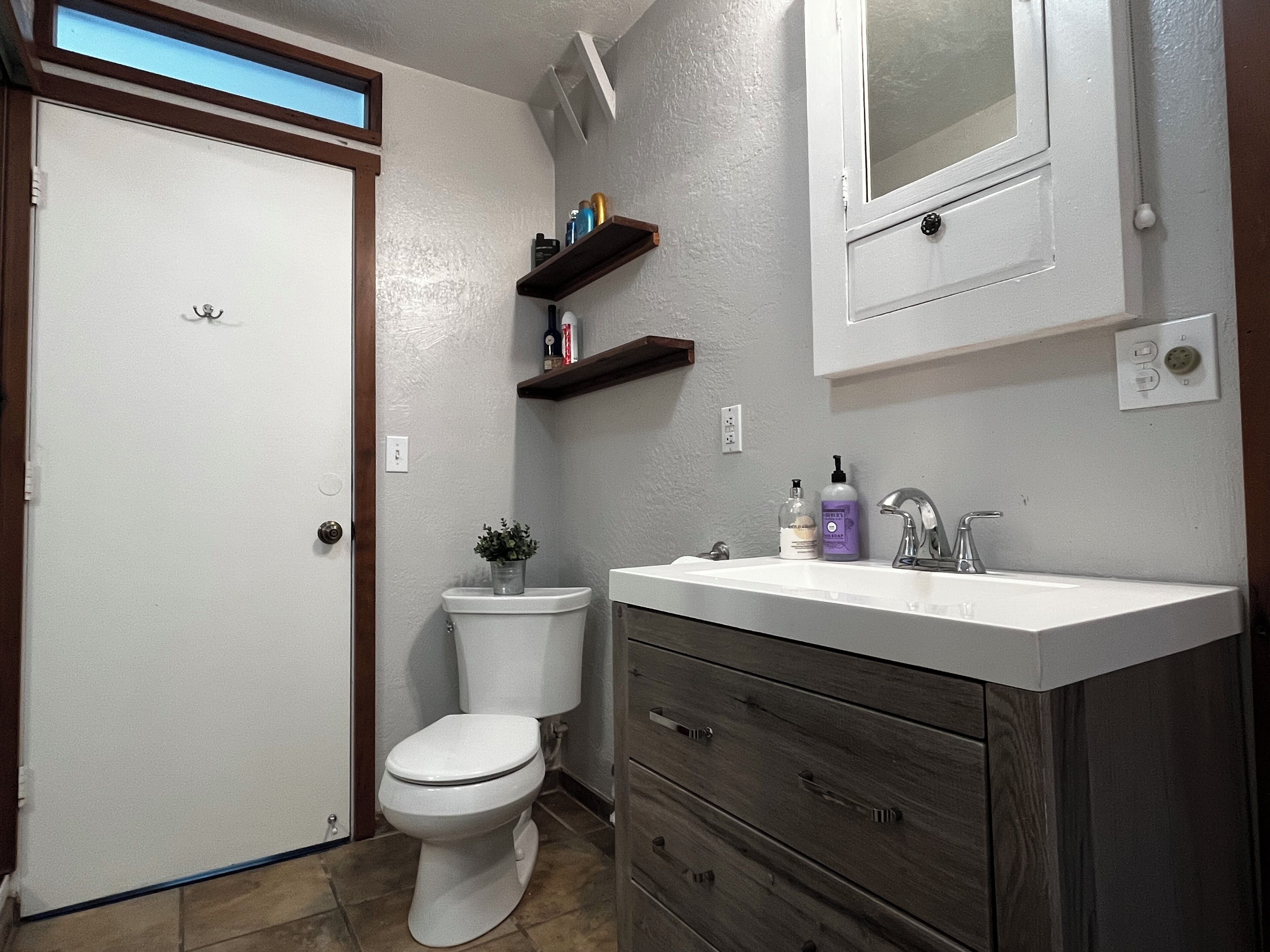 Un baño con inodoro, lavamanos y espejo encima. Hay repisas sobre el lavamanos con artículos de aseo.