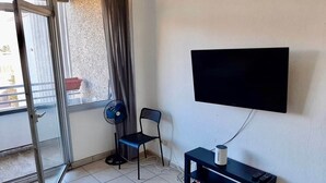 Een kamer met een schuifdeur, een zwarte stoel, een ventilator, een klein tafeltje met een afstandsbediening en een tv aan de muur.