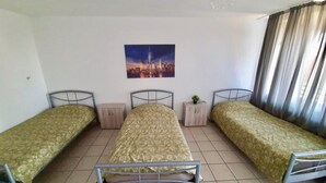 Een kamer met drie eenpersoonsbedden, een klein houten kastje, een potplant en een stadslandschap schilderij aan de muur.