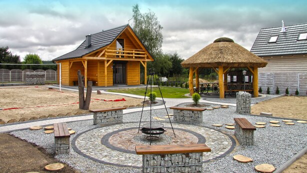 Una baita di legno con gazebo a tetto di paglia, circondata da un patio di ghiaia e pietra con panche e un braciere a forma circolare.