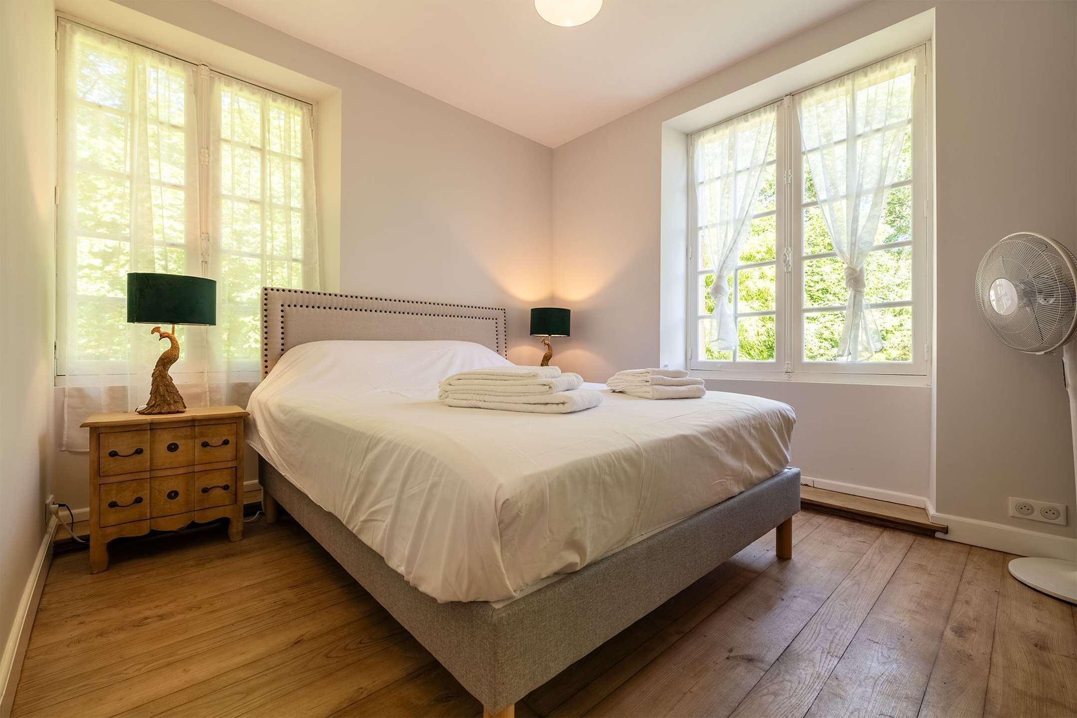 Une chambre à coucher avec un lit, deux tables de chevet avec des lampes, un ventilateur sur pied et de grandes fenêtres avec des rideaux.