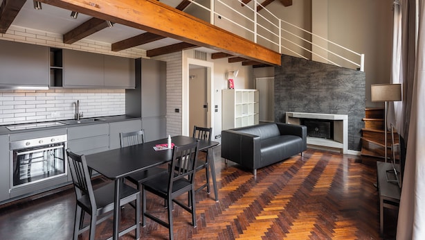 Une cuisine moderne avec un parquet en bois disposé en chevrons, un coin repas avec une table et des chaises noires, et un salon avec un canapé noir et une cheminée.
