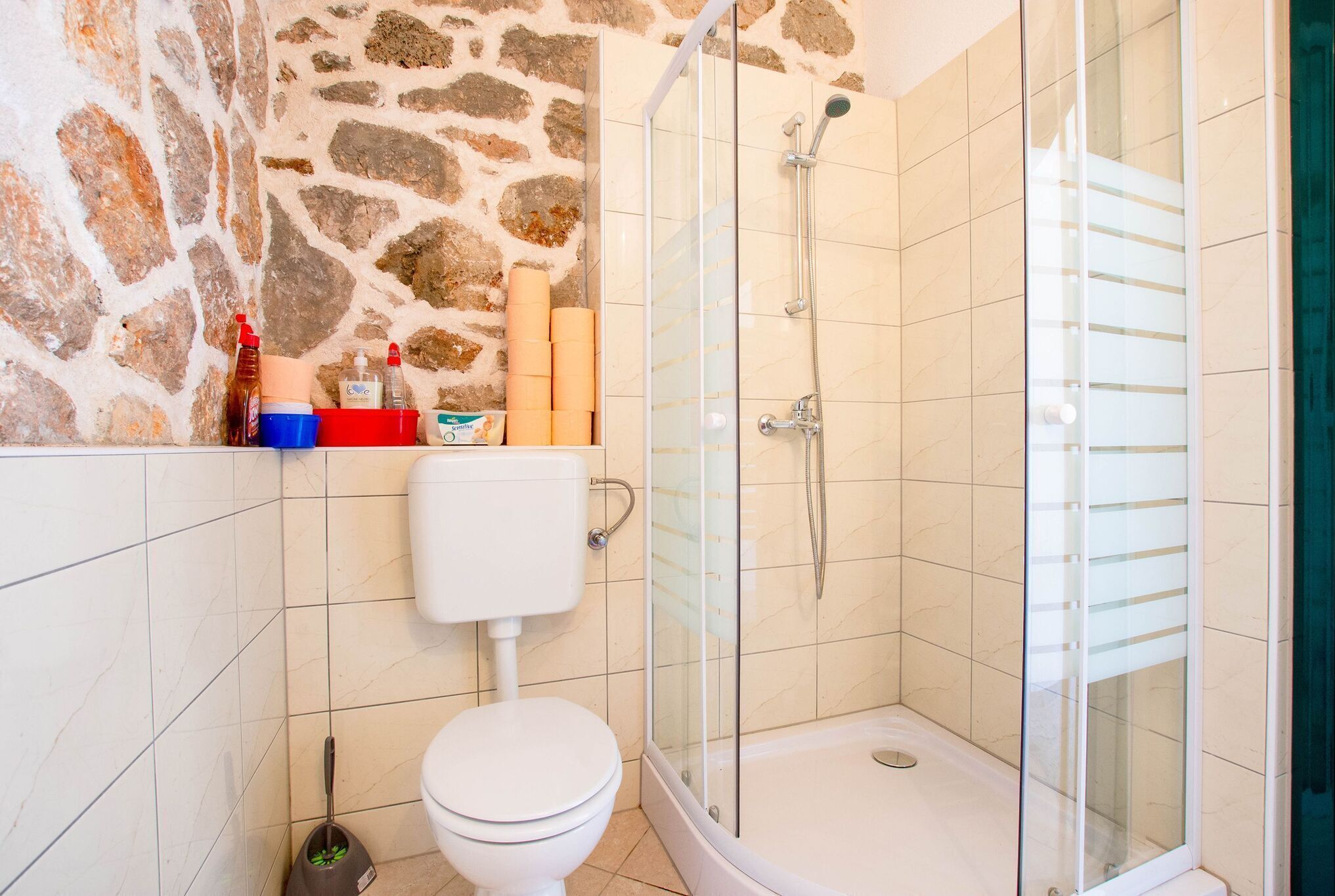 Een badkamer met een stenen muur, toilet, douche met glazen deur en een plank met toiletartikelen.