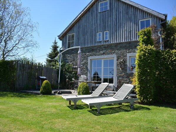 Une maison à deux étages avec une façade en pierre, une terrasse en bois et deux chaises longues.