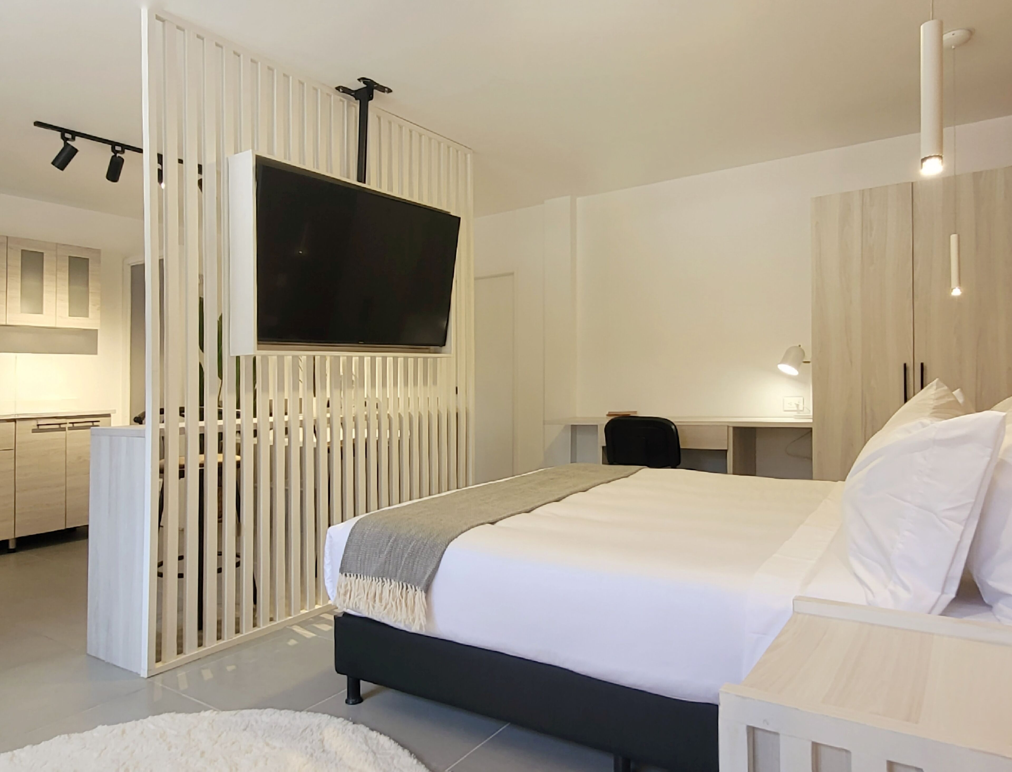 A modern hotel room with a large bed, a TV mounted on a white panel, a desk with a chair, and a small seating area.