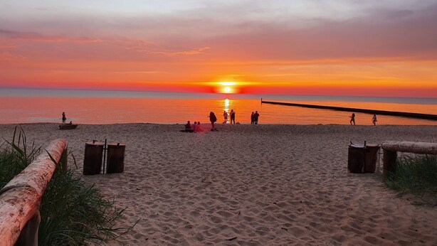 Ein Strand bei Sonnenuntergang, Menschen flanieren und sitzen im Sand.