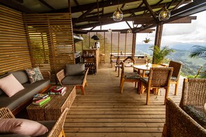 A wooden deck with wicker furniture, a sofa, and a dining area with a view of the mountains. - Owl's Watch Nature Retreat (Manizales)
