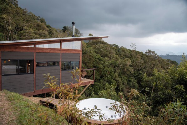 Una casa moderna con un jacuzzi en una terraza, rodeada de un bosque denso y colinas.