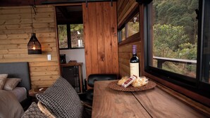 A cozy room with a wooden interior, a dining table with a bottle of wine and snacks, and a view of greenery outside. - Owl's Watch Nature Retreat (Manizales)