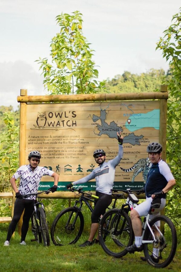 Three cyclists standing in front of a signboard with a map and text about a nature retreat and conservation project. - Owl's Watch Nature Retreat (Manizales)