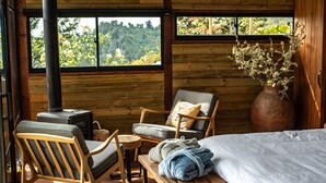 A wooden cabin interior with a bed, two chairs, a small table, a vase with dried flowers, and a wood-burning stove. - Owl's Watch Nature Retreat (Manizales)