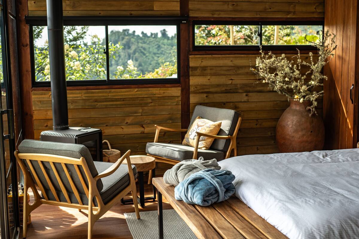 A wooden cabin interior with a bed, two chairs, a small table, a vase with dried flowers, and a wood-burning stove.