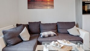 Moderni olohuone, jossa on kulmasohva, lasinen sohvapöytä ja seinälle ripustettu taulu, jossa on kuvattuna auringonlasku meren yllä.